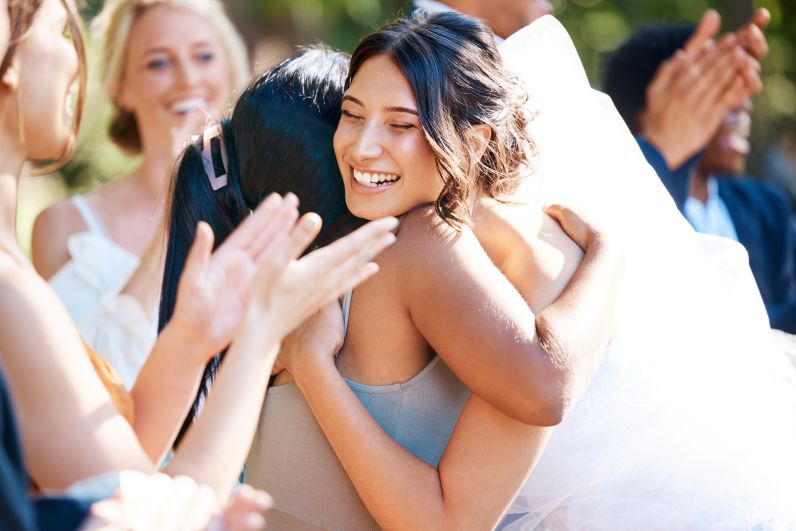Bride hugs a friend after the wedding ceremony as guests celebrate in the background.