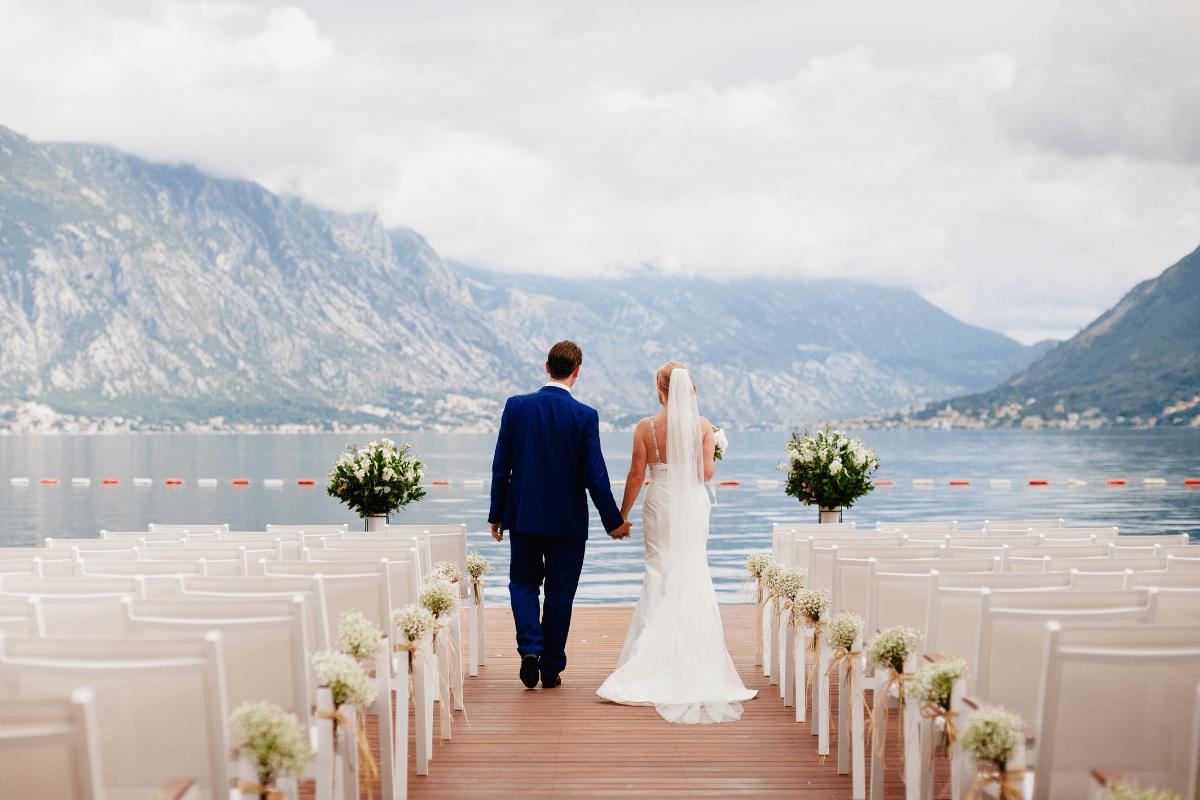 Couple walks hand in hand down an outdoor wedding aisle overlooking a lake and mountains.