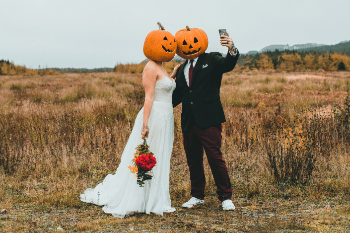 Halloween wedding couple wearing jack-o'-lantern pumpkin heads and taking a selfie outdoors