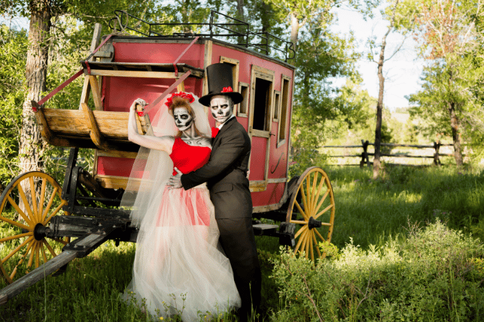 Halloween wedding bride and groom in skeleton makeup posing beside a vintage carriage