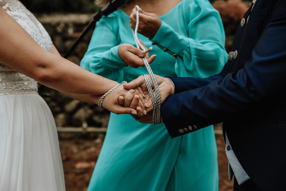 Couple holding hands as an officiant wraps a cord around their wrists during a handfasting ceremony