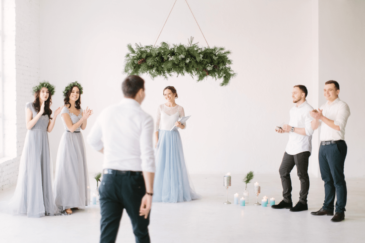 rehearsal.png Wedding party rehearses an indoor ceremony beneath a greenery arch.