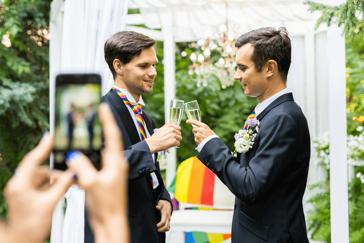 Two grooms toast with champagne at an outdoor wedding as a guest takes their photo.