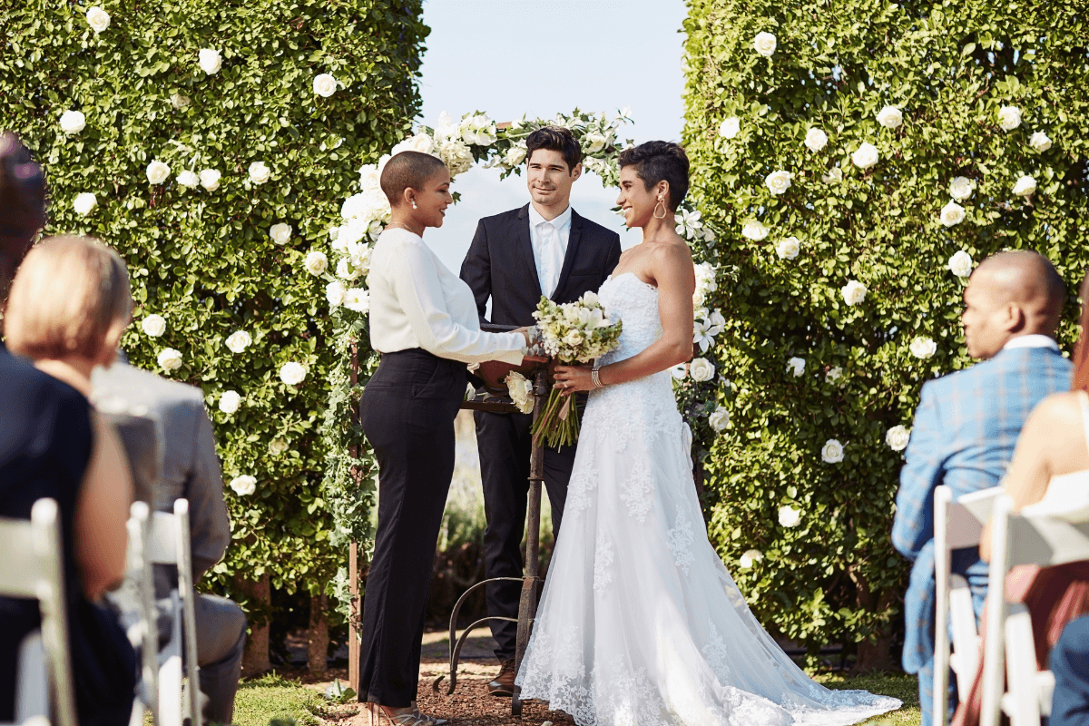 Wedding officiant leads an outdoor ceremony as the couple stands together beneath a floral backdrop.