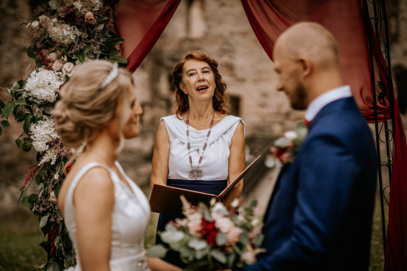 Wedding officiant leads an outdoor wedding ceremony as the couple exchange vows beneath a floral arch.
