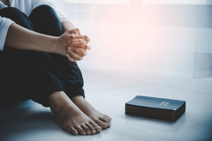 Person sitting on the floor with hands clasped beside a Bible