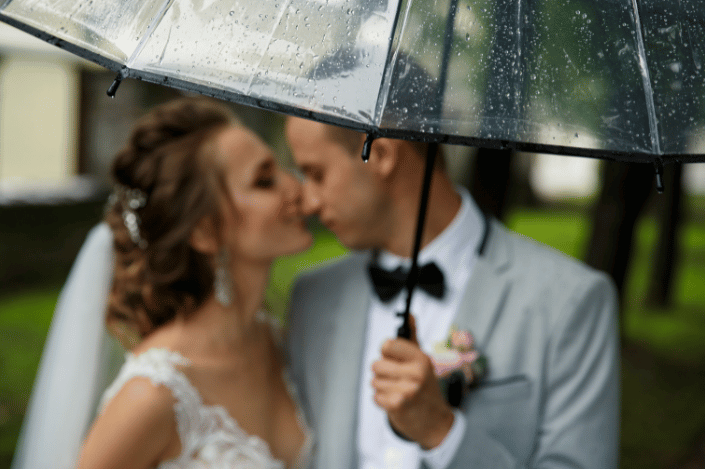 Bride and groom sharing a kiss under a clear umbrella on a rainy wedding day.