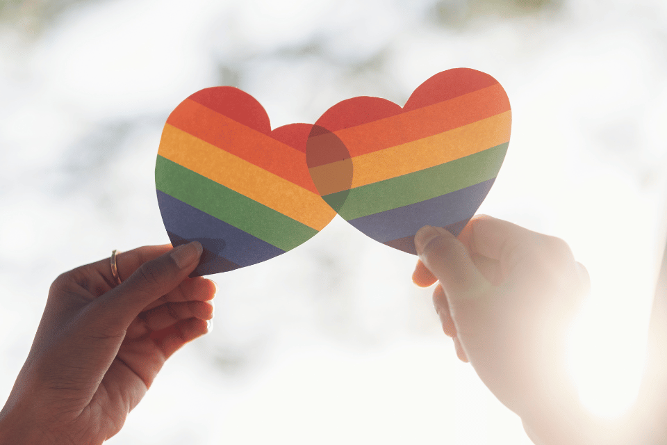 Two hands holding overlapping rainbow hearts representing LGBTQ love and inclusion