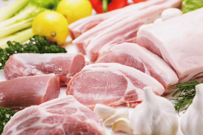 Assorted raw pork cuts displayed with fresh herbs and vegetables, illustrating foods prohibited by some religions.
