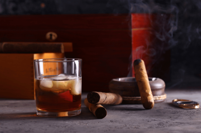 Glass of whiskey beside cigars and a smoking cigar in an ashtray, illustrating religious restrictions on intoxicants.