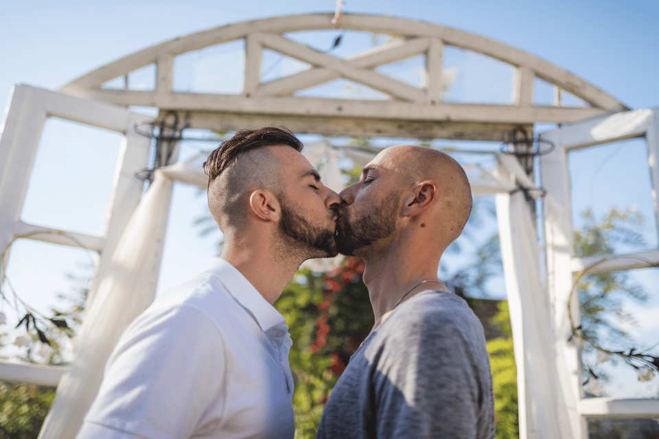 Two grooms kissing beneath an outdoor wedding arch