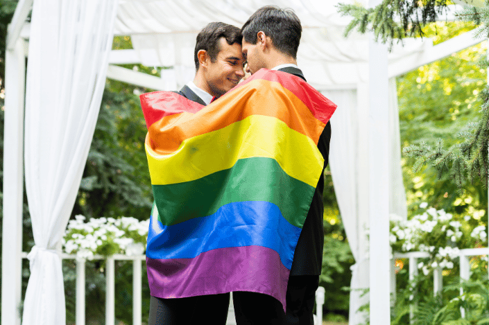 Two grooms embracing under a rainbow flag at an outdoor wedding