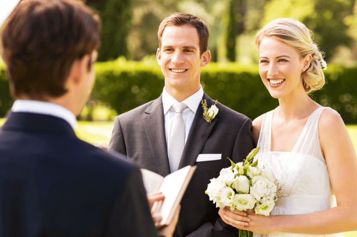 Wedding officiant leading a ceremony for a smiling couple outdoors.