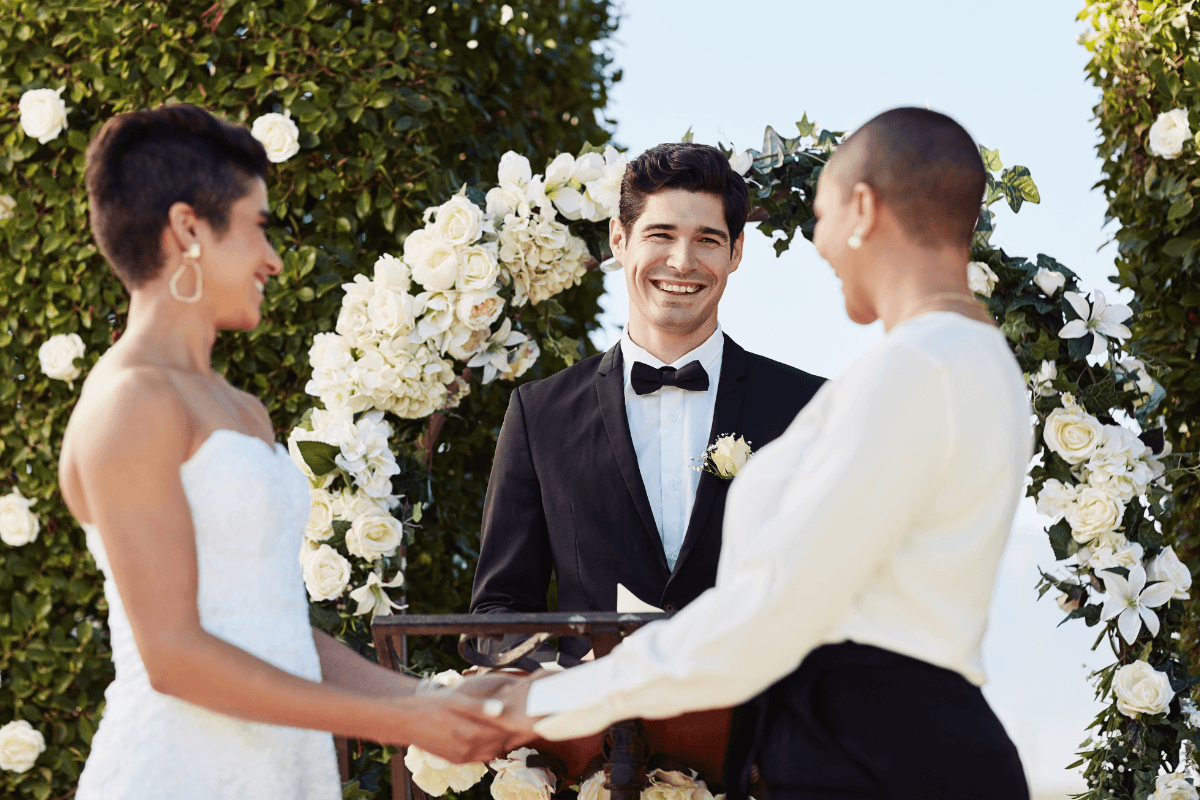 Wedding officiant smiles as a couple holds hands during an outdoor ceremony beneath a white floral arch.