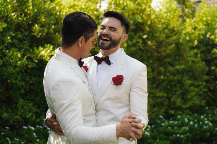 Two grooms in white suits smiling and dancing together outdoors