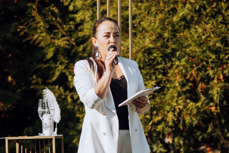 Wedding officiant speaks into a microphone while leading an outdoor wedding ceremony beneath a floral arch.