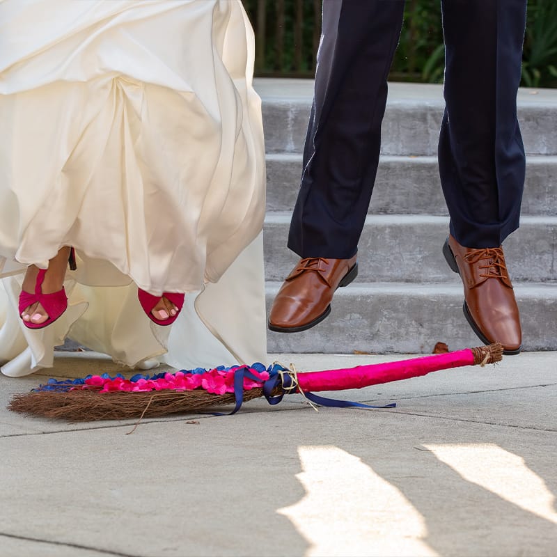 A newlywed couple jumping over a broom that's been decorated.